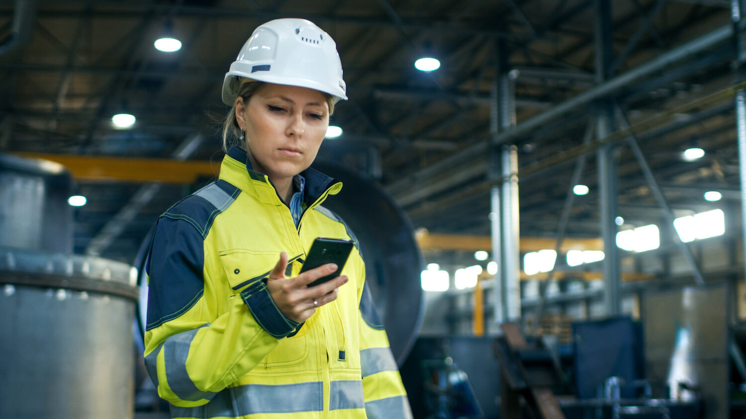 Female Industrial Worker in the Hard Hat Uses Mobile Phone While Walking Through Heavy Industry Manufacturing Factory