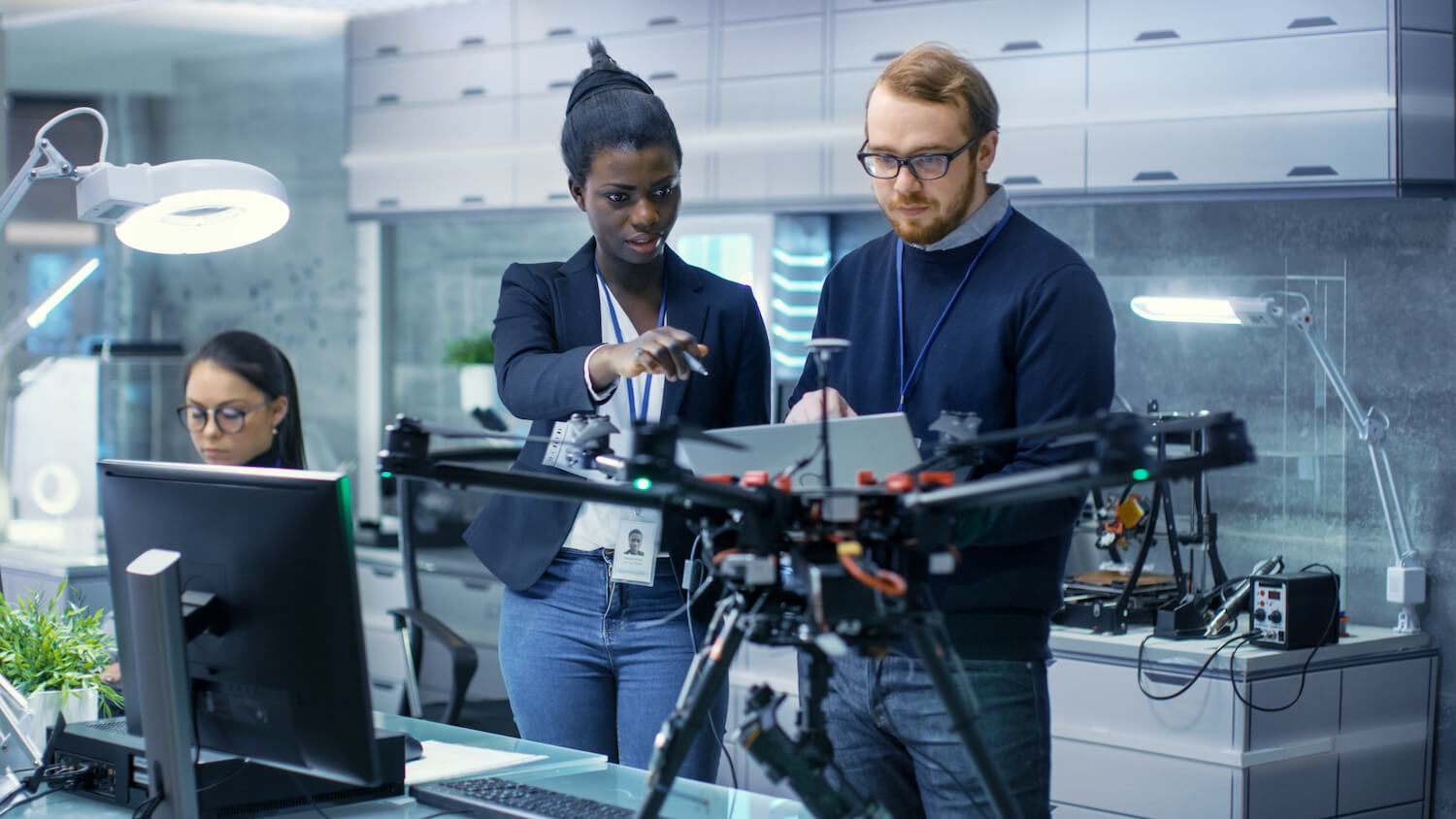Caucasian Male and Black Female Engineers Working on a Drone Project with Help of Laptop and Taking Notes